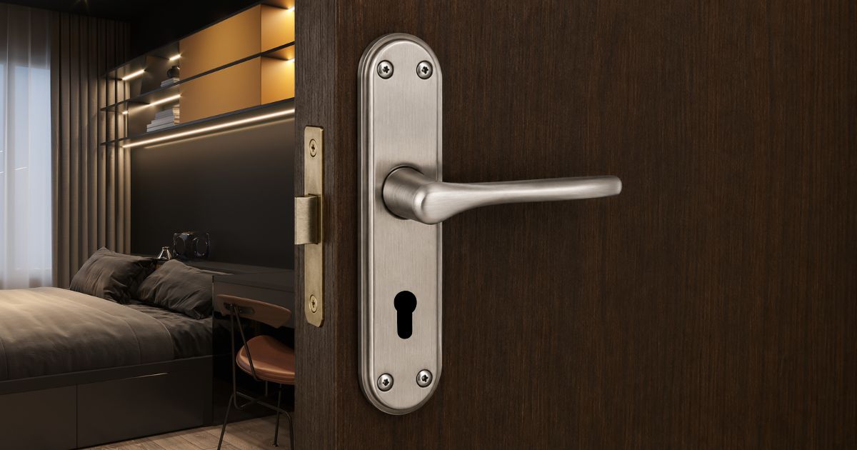 Close-up of a brushed metal door handle and keyhole plate on a dark wood door, with a modern bedroom visible in the background.