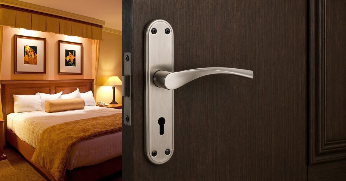 Hotel room visible through an open dark wooden door with a silver lever handle, featuring a king bed, lamps, and framed artwork.