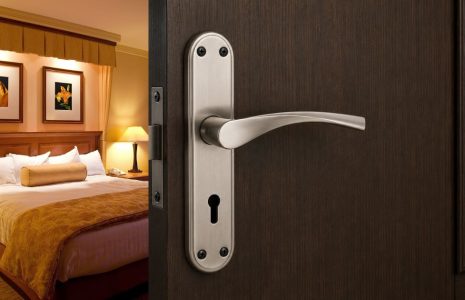 Hotel room visible through an open dark wooden door with a silver lever handle, featuring a king bed, lamps, and framed artwork.